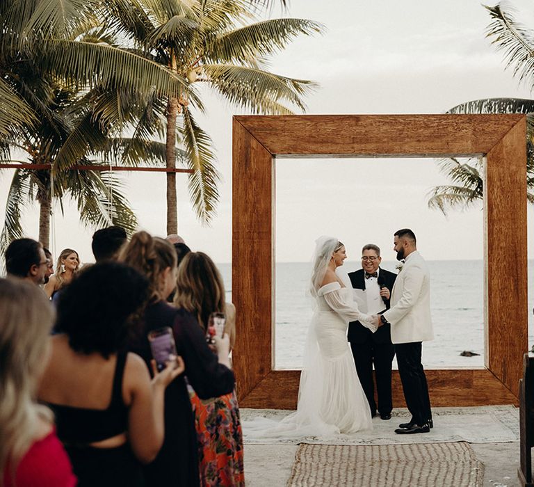 Bride in a strapless tulle wedding dress and groom in a white tuxedo jacket exchanging vows by the ocean at their blue venado beach wedding ceremony 