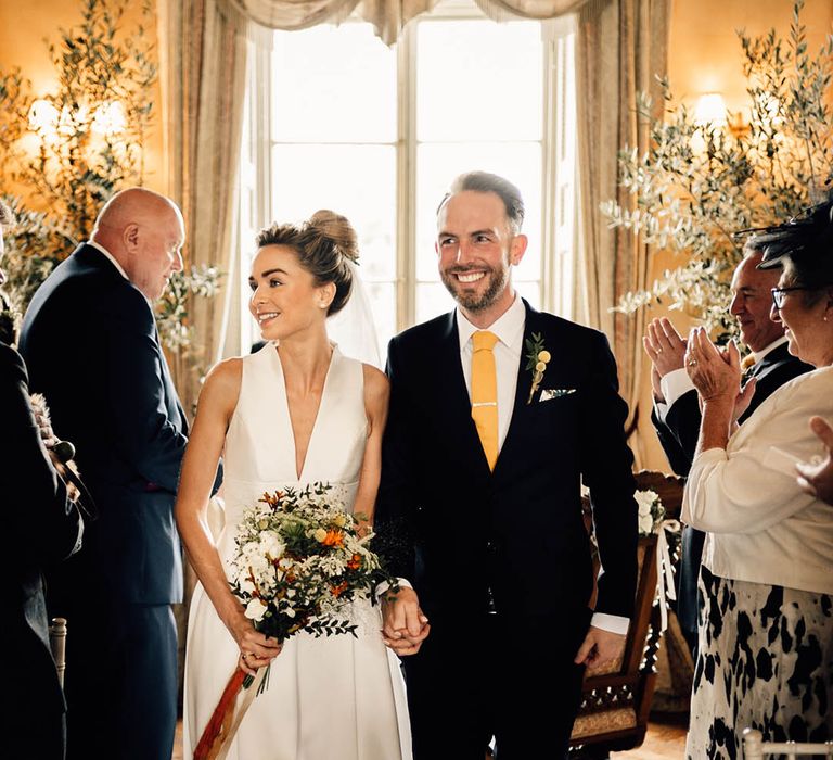 Groom in navy suit with yellow tie and tie clip walking back down the aisle with the bride in a plunging wedding dress carrying an orange bouquet 