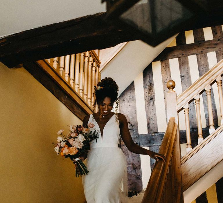 Bride in v-neck sleeveless wedding dress with 3D floral applique train holding blush and orange garden roses, poppies and carnations, dried flowers and eucalyptus wedding bouquet walking down the stairs at Brinsop Court wedding venue