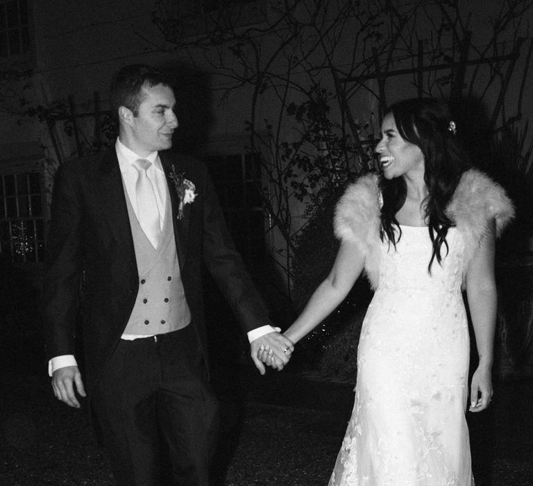 Groom in navy grooms morning suit with grey waistcoat, off-white tie and mixed wildflower boutonniere walking with bride in a-line tulle wedding dress with beaded lace applique with petals and a square neckline with side tulle cutouts and faux fur bridal coverup at South Farms wedding