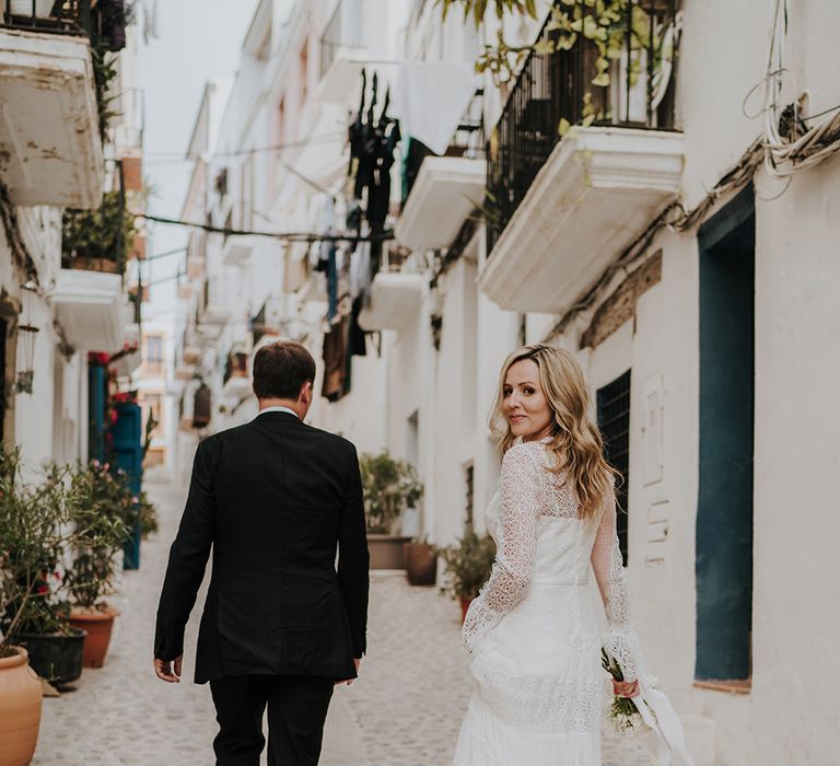 bride in a boho lace wedding dress with long sleeves walking through the side streets of ibiza with white buildings balconies and shutters with her groom in a black suit