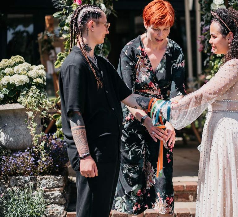 Wedding celebrant in wrap around black and floral patterned dress leading handfasting ritual at at-home ceremony for bride in long sleeve v-neck sheer polka dot overlay sparkly wedding dress and gold celestial bridal headband and bride in black shirt and black trousers at Viking inspired wedding