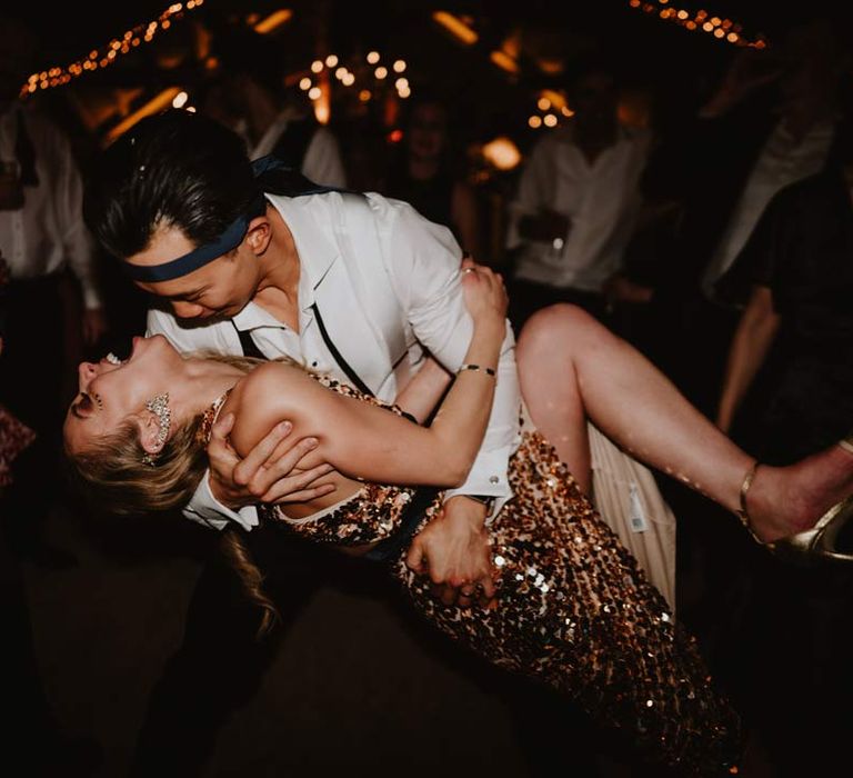 Groom in white shirt with black tie around his head dancing and laughing with bride in gold sequin sparkly reception dress 