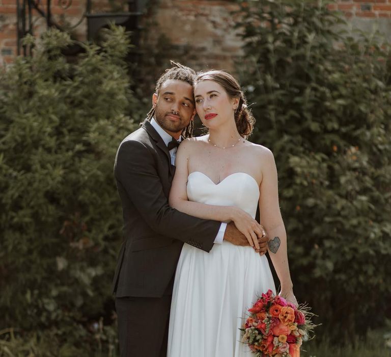 Bride in strapless sweetheart neckline wedding dress holding pink and orange wedding bouquet being embraced by groom in classic black tuxedo with bowtie and pink and orange wedding boutonniere at The Old Rectory Berkshire