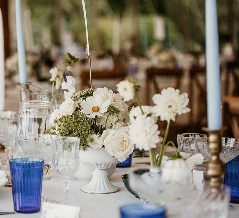 White cosmos, dahlias, and roses in white bowl table centrepiece with blue coloured glassware 