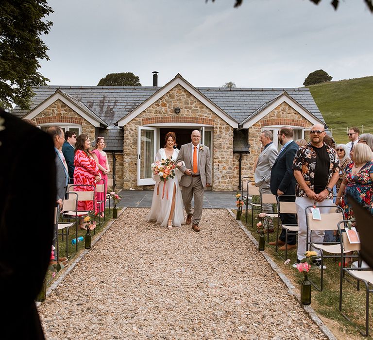 Father of the bride wearing orange colourful wedding tie walks the bride down the aisle at outdoor wedding 