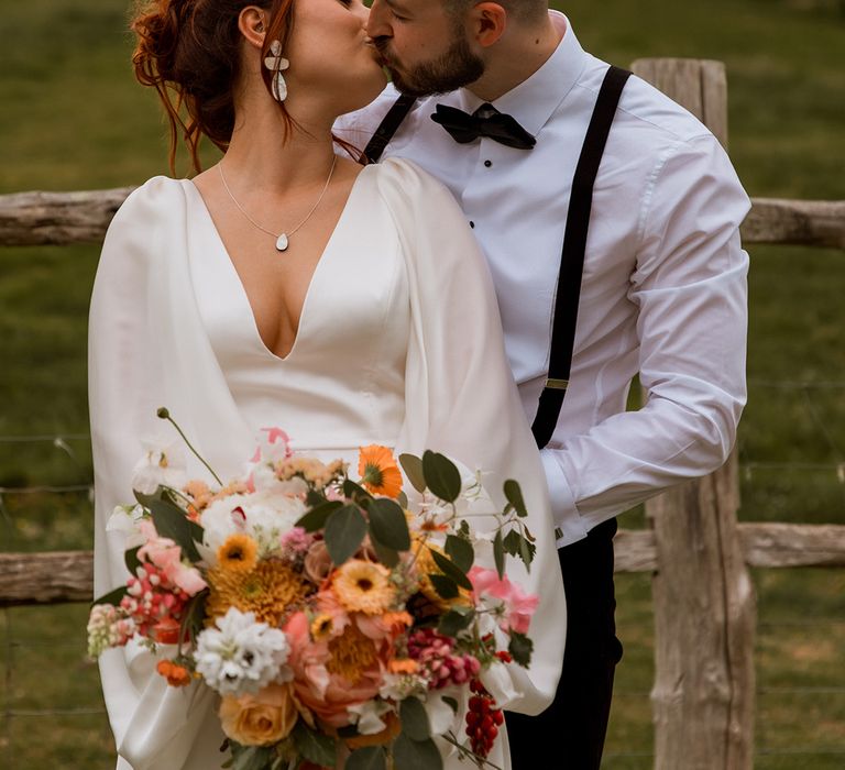 The bride in a gorgeous satin gown holding orange wedding bouquet kissing groom in black tie 