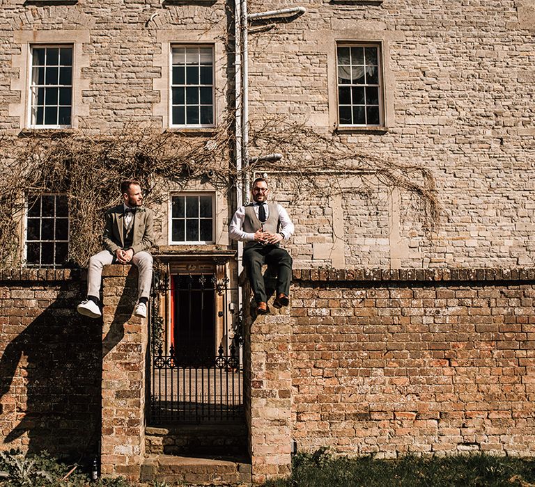 Two grooms sitting on a high brick wall next to a gate at country house wedding venue in Gloucester 