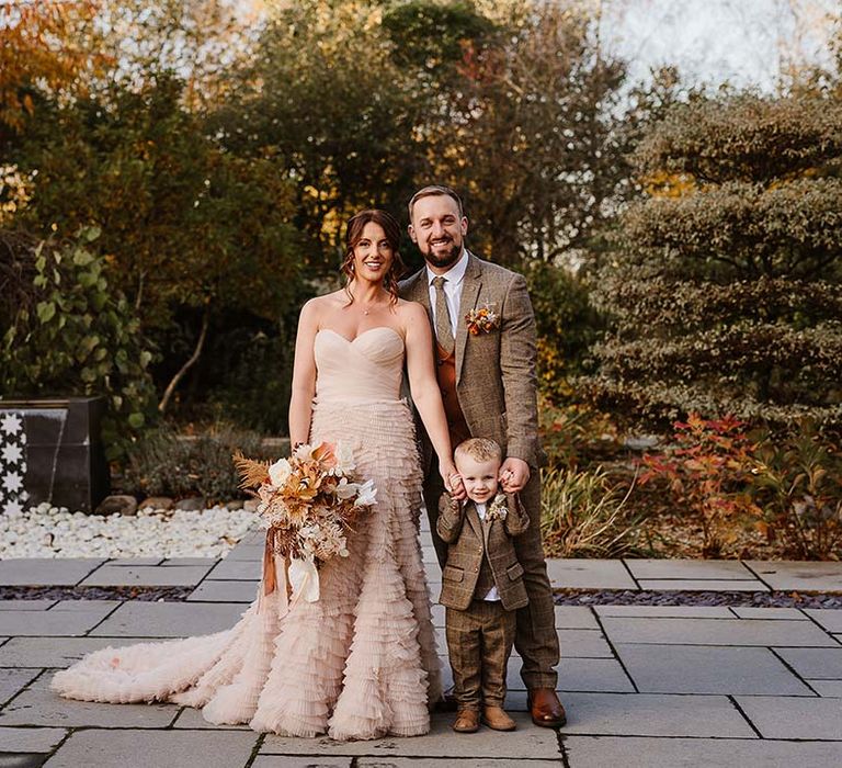 The bride wears a strapless ruffle wedding dress from Recycle My Wedding with the groom in a brown wedding suit with their son in a matching suit 