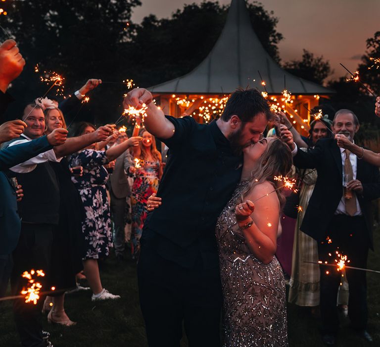 The bride and groom in their wedding reception outfits share a kiss at their sparkler send off 