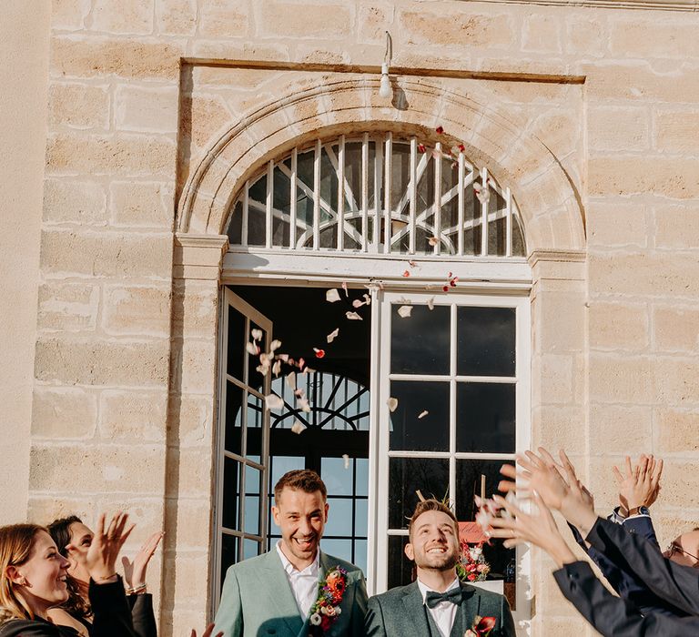 Two grooms walk out of their chateau wedding to confetti thrown by their guests 