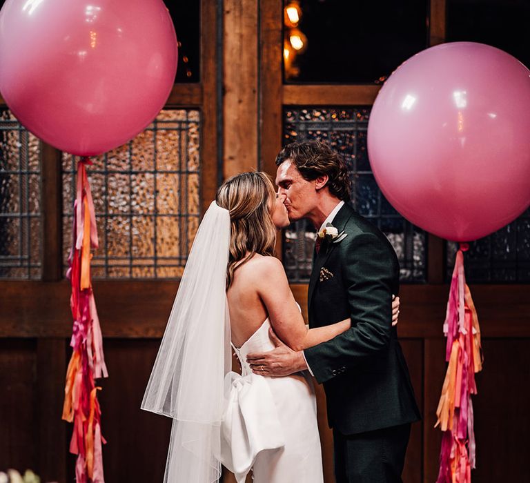 First kiss for the bride and groom at their wedding ceremony with large pink balloons 