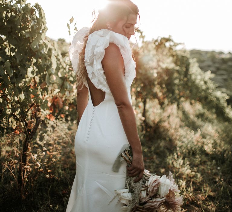 Bride in a Rime Arodaky wedding dress with low back detail, buttons and ruffle sleeves standing in a field 