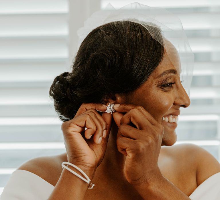 Black bride with side pinned hair and blush veil putting on her earrings 