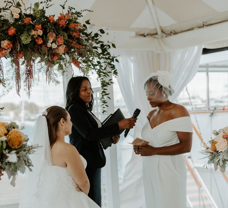 Emotional black bride in an off the shoulder jumpsuit and blush veil reads her vows to her bride at same sex wedding 