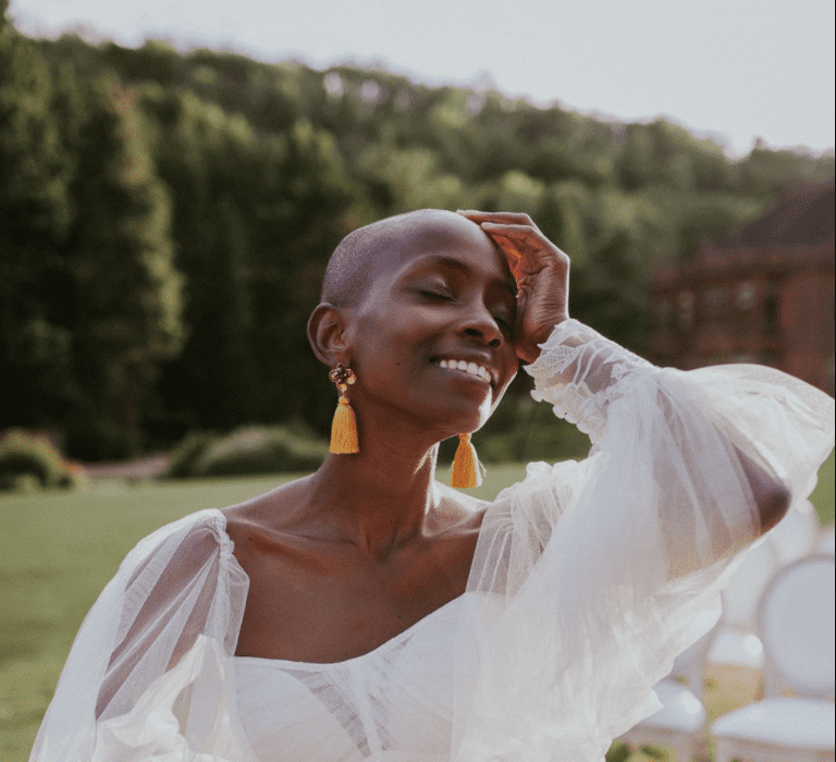 A Black bride with a shaved head looks towards the sky with a hand loosely to her forehead. She wears statement yellow earrings and a wedding gown with floaty sleeves.