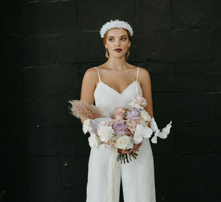 Stylish bride in a sequin jumpsuit with thin straps holding a pastel pink and purple wedding bouquet 