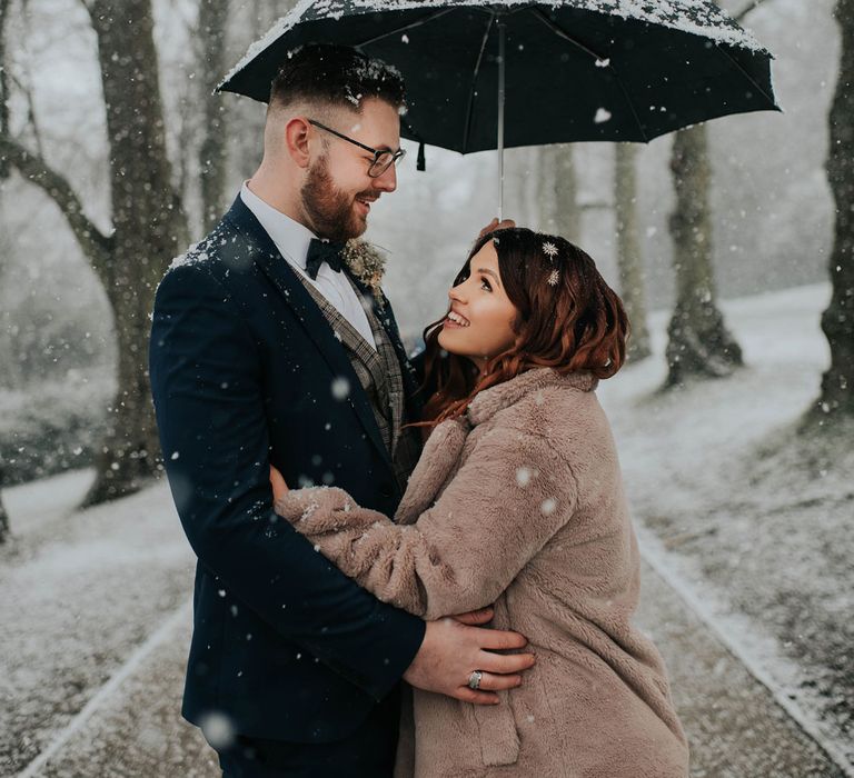 Bride in fur coat smiles up at groom in navy suit whilst both stand under a black umbrella during snowfall at Cannon Hall