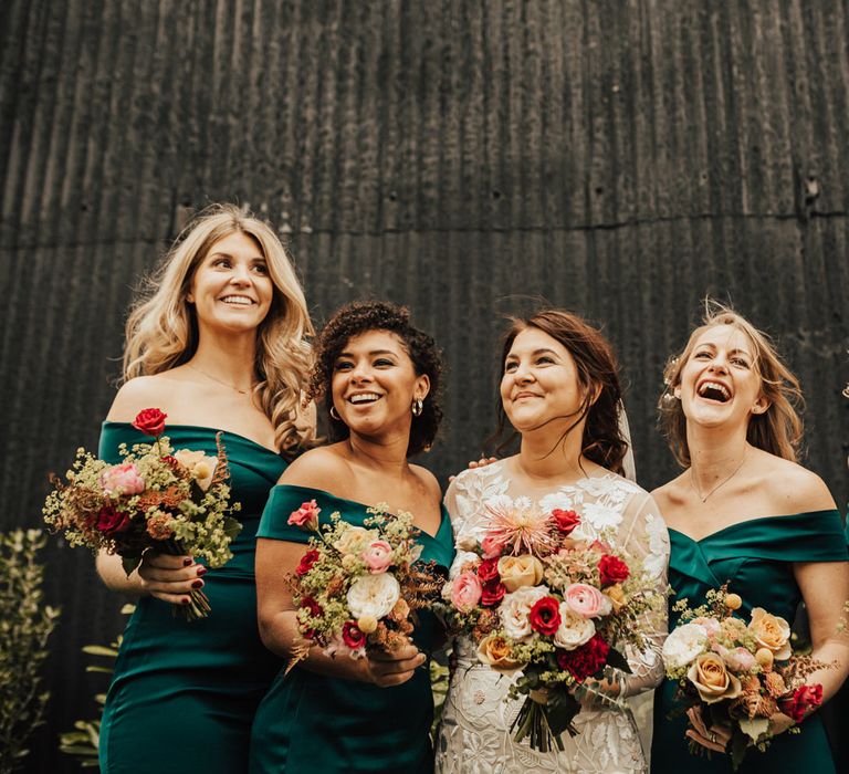 Bridal party portrait with bride in a lace wedding dress and bridesmaids in off the shoulder dark green dresses holding red, orange and yellow flower bouquets 