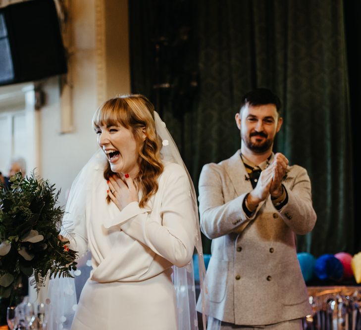 Smiley bride in a jumpsuit and appliqué veil holding a green foliage wedding bouquet with red nail polish