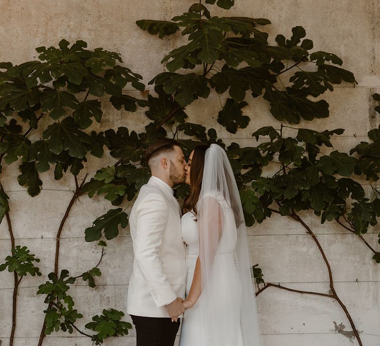 Bride in V neck Made With Love wedding dress and chapel length veil stands holding hands with groom in white double breasted tuxedo jacket