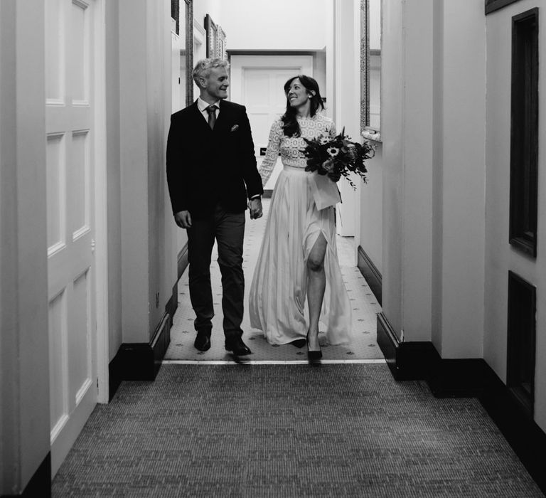 Bride in long sleeved lace top Self Portrait wedding dress holding colourful bouquet walks down Bristol Registry Office hallway holding hands with groom in brown woollen blazer and waistcoat