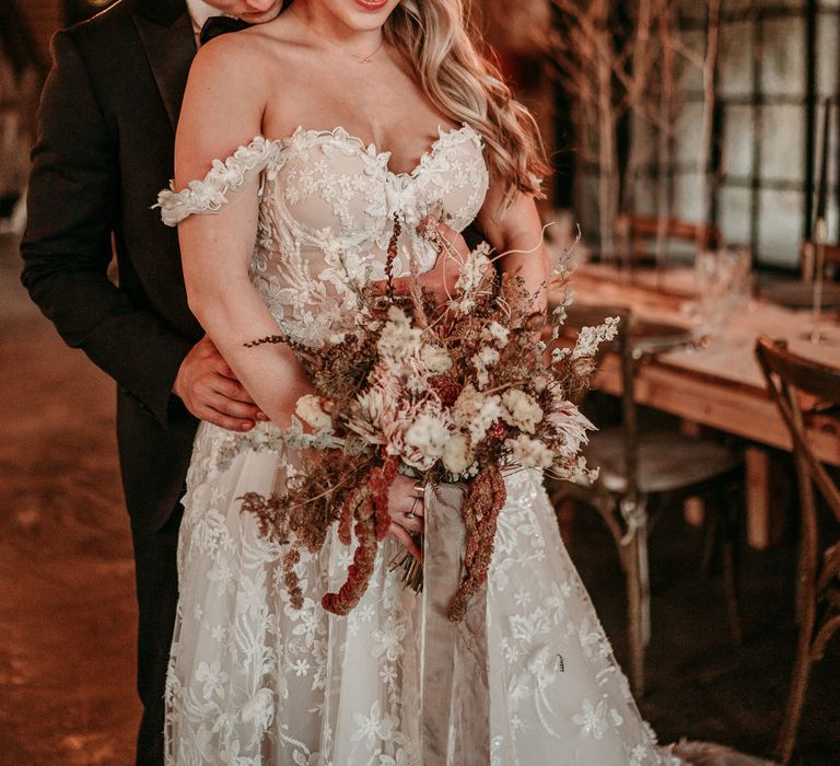 Groom in a black tuxedo embracing his bride in an off the shoulder appliqué wedding dress at The Hidden River Cabins