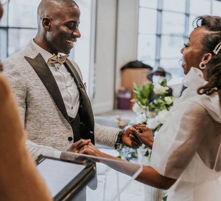 Laughing bride in white Bec + Bridge wedding dress with shoulder bows holds hands with groom in grey Moss Bros suit jacket at the altar during Bridge Community Church Wedding