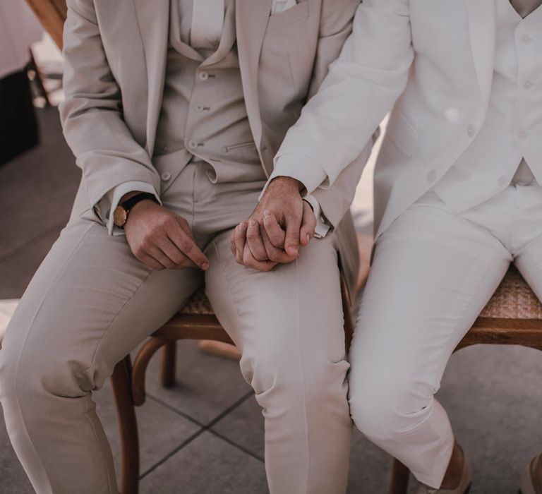 Grooms in light grey wedding suit and white wedding suit hold hands during ceremony