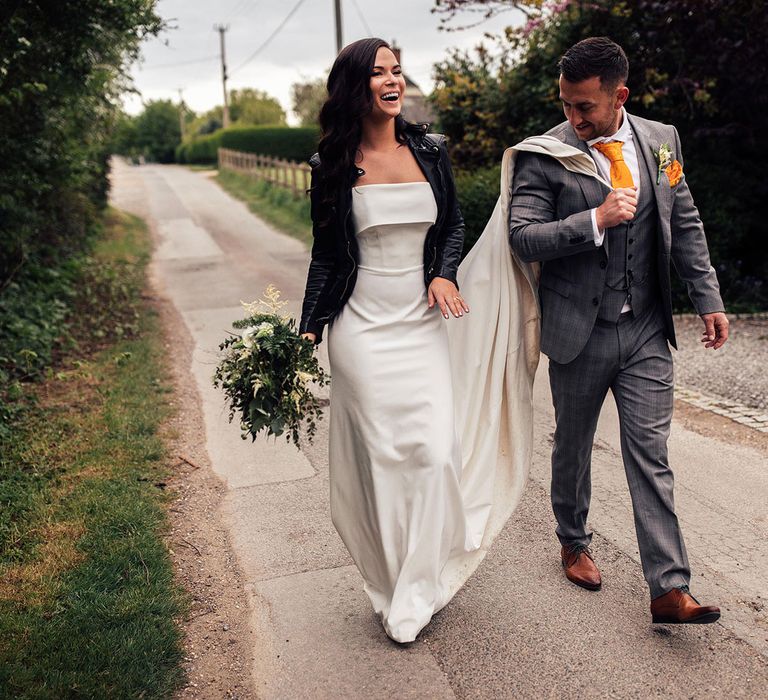 Bride in white Vagabond wedding dress and bridal leather jacket walks with groom in grey Hugo Boss suit with yellow tie as he holds her train