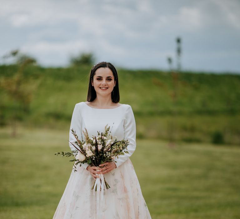 Beautiful bride with bobbed hair in a couture wedding dress with long sleeves and floral patterned skirt holding a pink and white peony bouquet 