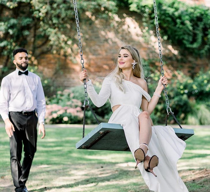 Stylish bride in a one shoulder wedding dress with long sleeve and black sandals sitting on a swing at Garthmyl Hall 