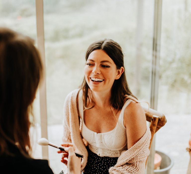 Laughing bride with long brown hair, white cami top and beige cardigan gets ready for her wedding