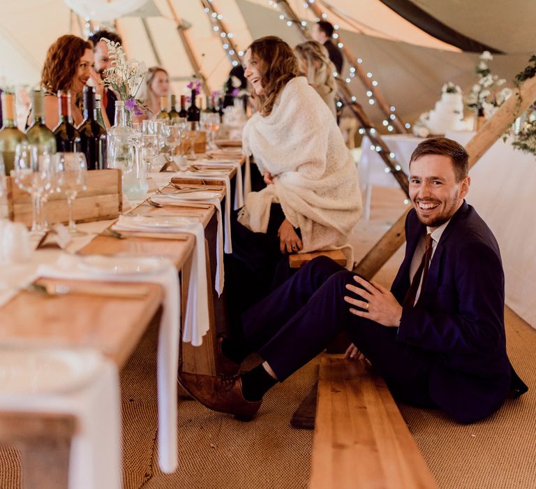 Wedding guest in dark blue suit sits on the ground after falling down bench inside tipi wedding reception with fairy lights, foliage decoration and rustic table at home farm wedding