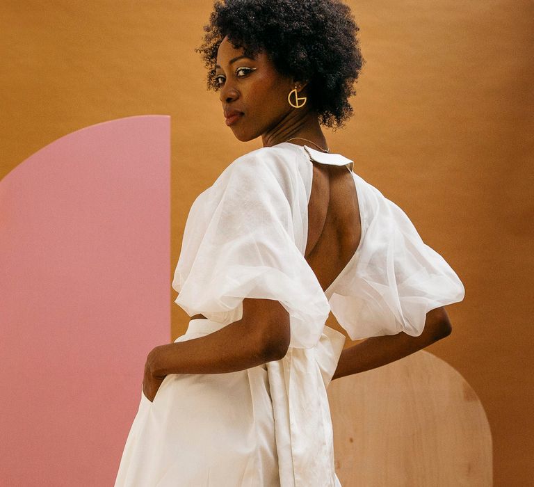 Bride with afro hair wearing puff sleeve, open back wedding separates with bow detail in front of a mustard colour backdrop