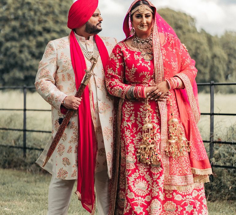 Bride & groom stand beside one another as bride wears traditional Lengha in red and gold 