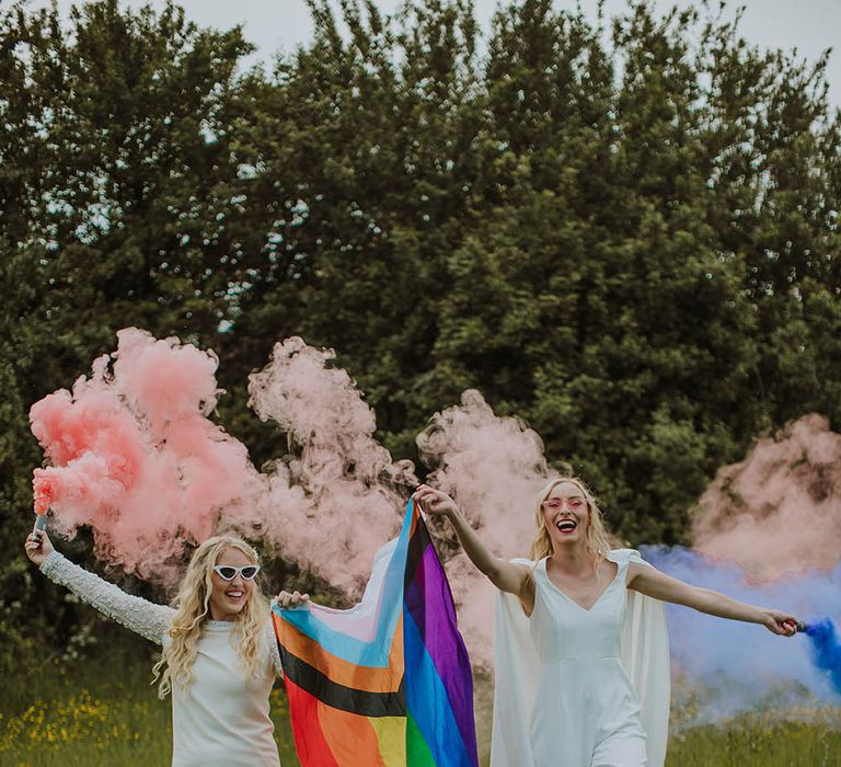 Two brides in a short wedding dress with long sleeves and bride in a jumpsuit and cape holding smoke bombs and a pride flag 