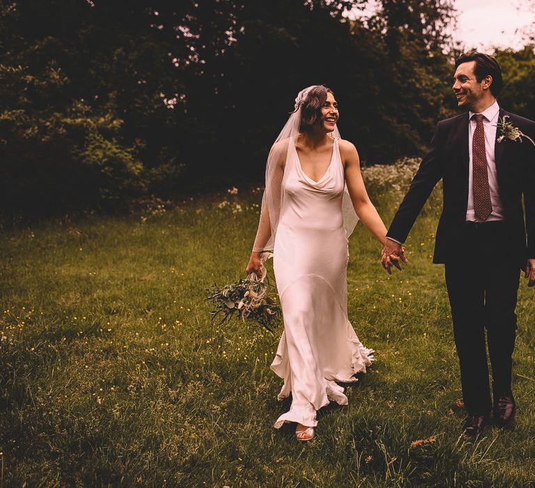 groom in a black suit with red tie holding hands with his bride in a satin wedding dress with cowl front and Juliet cap wedding veil in a field