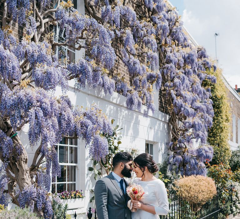 Bride & groom stand in front of hanging purple wisteria tree within Chelsea on their wedding day