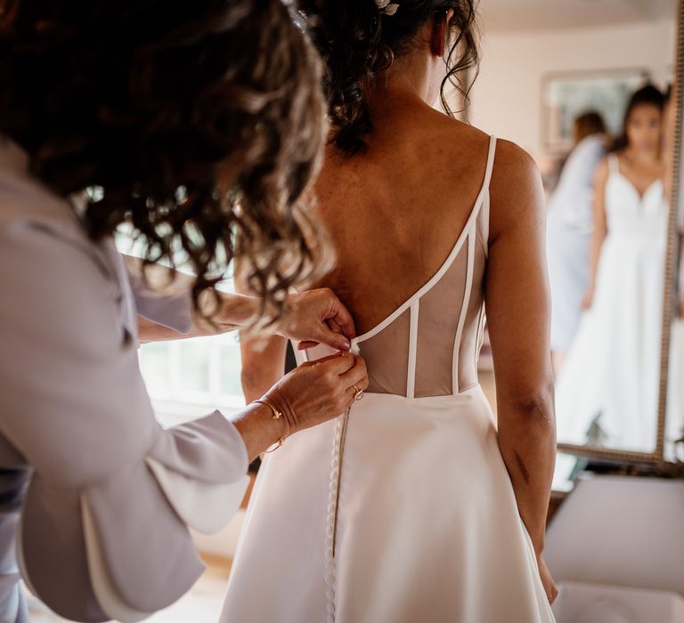 Bride in low mesh back wedding dress with button detailing gets her buttons fastened by mother in light purple dress before wedding at The West Mill Derby