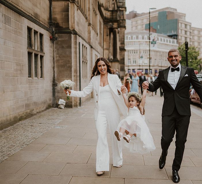 Family wedding portrait with bride and groom in black and white suits swinging their flower girl daughter in the air 