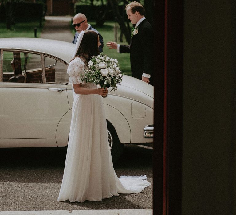 Bride in lace puffed sleeved Daalarna wedding dress holding white and green bridal bouquet stands in front of white classic car outside church before wedding ceremony in Surrey