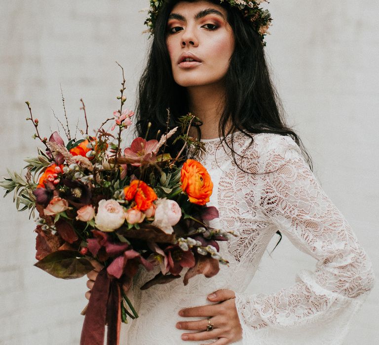 Stylish bride in a lace wedding dress with long bell sleeves holding an orange, and red wedding bouquet 