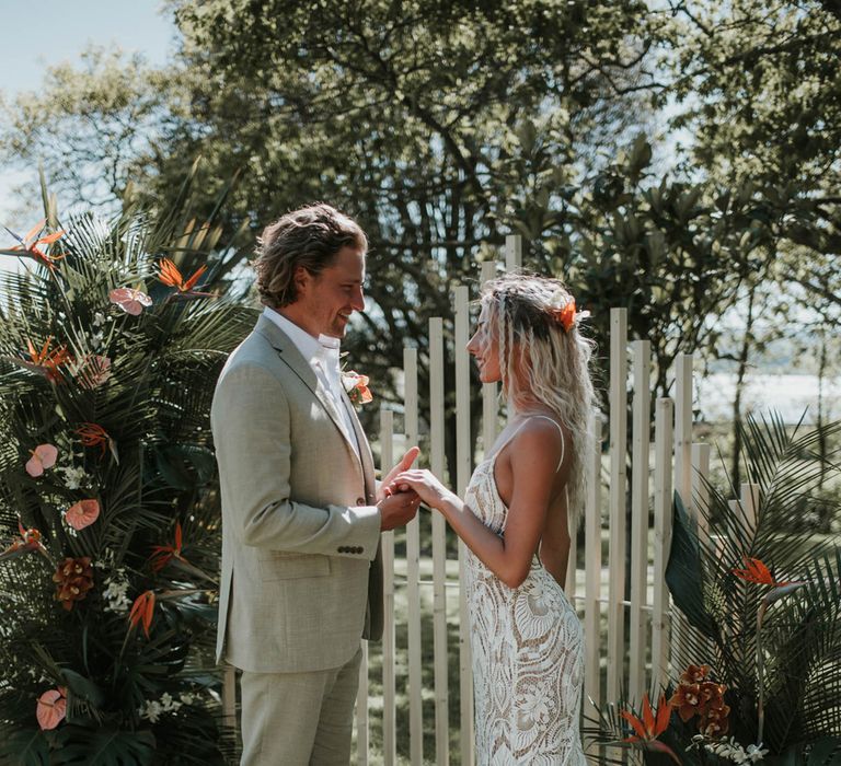 Groom in white shirt and linen suit holds hands of bride with blonde beach waves wearing lace low boho wedding with low back in front of tropical flower installation for birds of paradise wedding inspiration