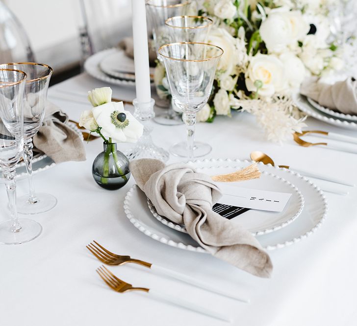 A black and white wedding place setting has two plates, a neutral coloured tied napkin, gold cutlery, place tag with gold tassel and tall white tapered candles. There is bud vases with lithianthus and anemones and a white table cloth