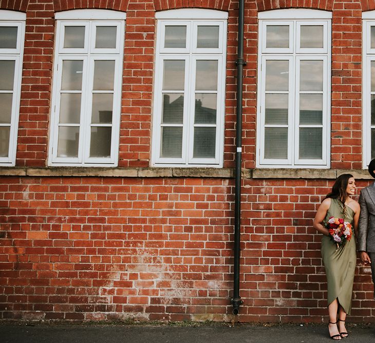 An Indian British couple stand against a brick wall for a wedding portrait. He wears a grey suit and she wears a d ark green dress and holds a colour pink, orange and red bouquet.