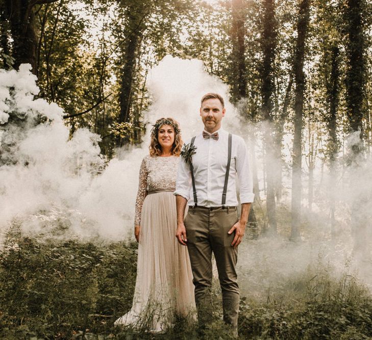 Woodland wedding with bride in a lace wedding dress and flower crown standing with her husband in braces and bow tie as a white smoke bomb goes off in the background 