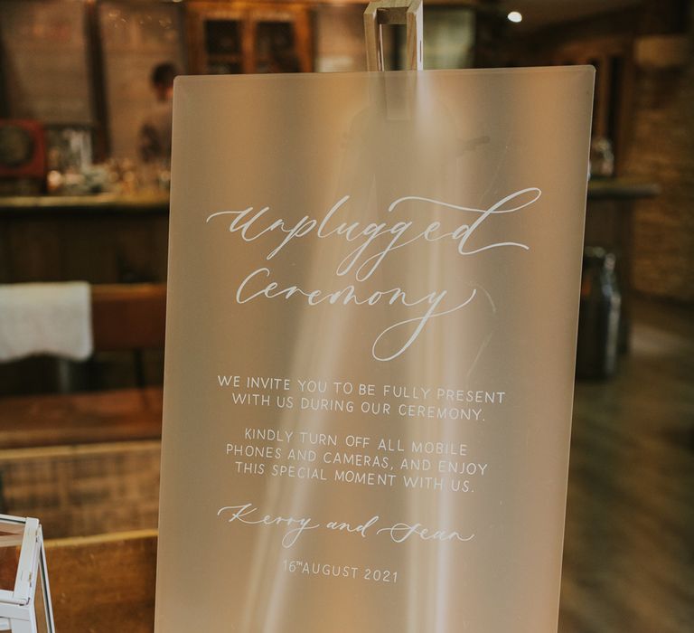 Frosted acrylic wedding sign reading 'unplugged ceremony' on wooden table with white flowers and green leaf decor for Tythe Barn wedding with barn wedding flowers