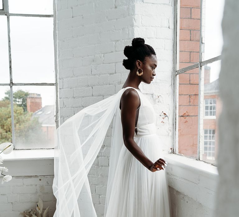 Black bride in a tulle skirt wedding dress standing at the window with a Watteau train attached to the shoulders of the wedding dress