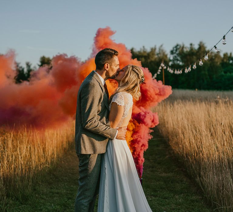 Bride in white lace top open back wedding dress with train kisses groom in grey herringbone suit as red and orange smoke bomb goes off behind them during late summer wedding at Wellington Wood Norfolk